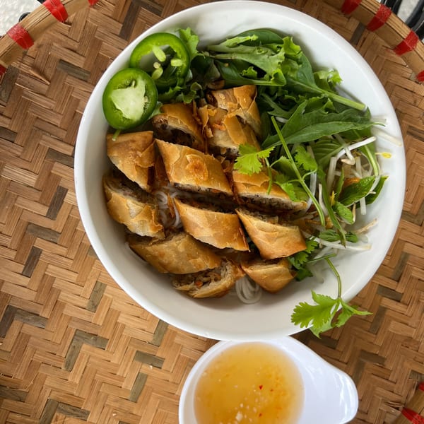 Vietnamese vermicelli bowl with crispy spring rolls, fresh herbs, and sliced jalapeños, with a side of sauce.